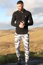 A man stands outdoors wearing a black Prestige Camo Hooded Tee and white, gray, and black camouflage cargo pants.